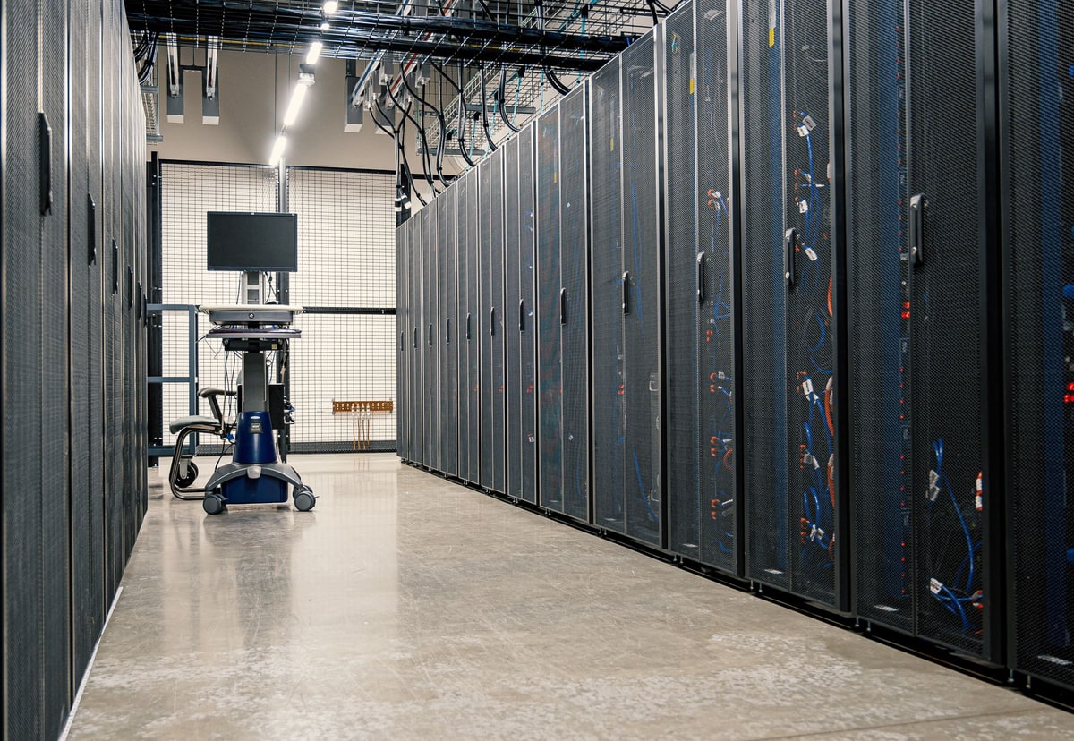 Rows of black server racks in a modern data centre corridor with visible cabling and maintenance equipment