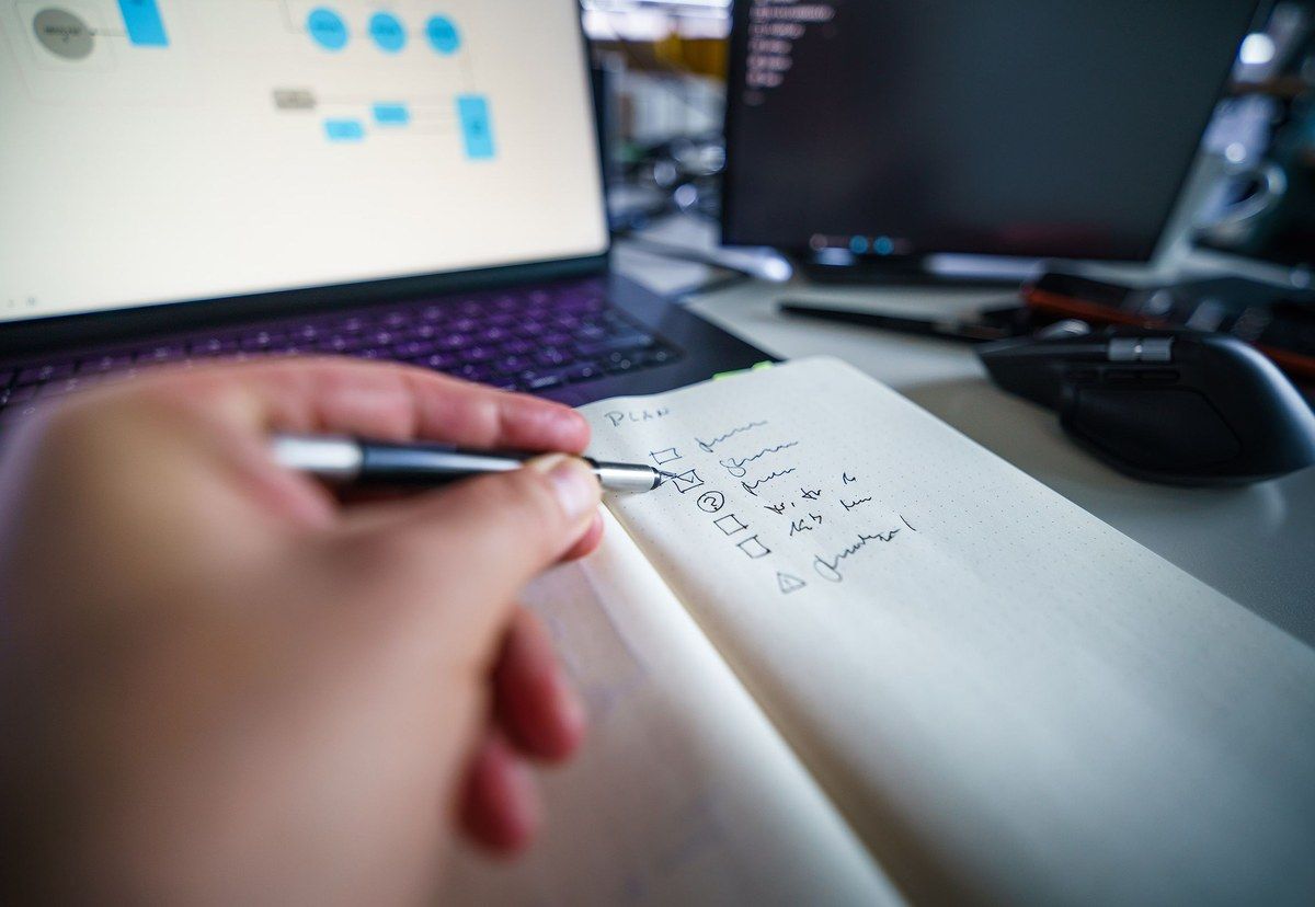 A person checks a handwritten task list beside a laptop and two work screens