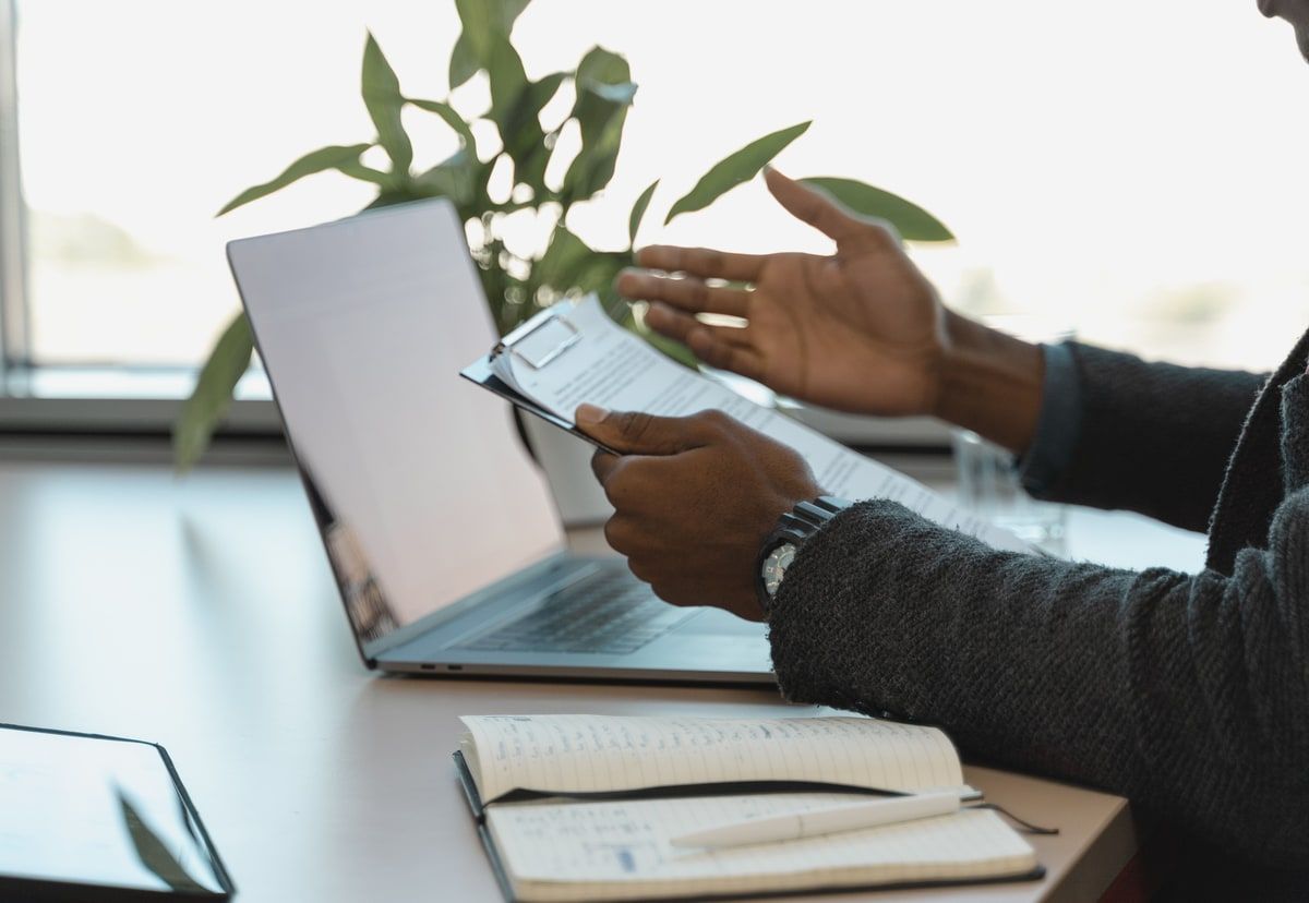 A business professional holds a clipboard and gestures beside an open laptop and notebook