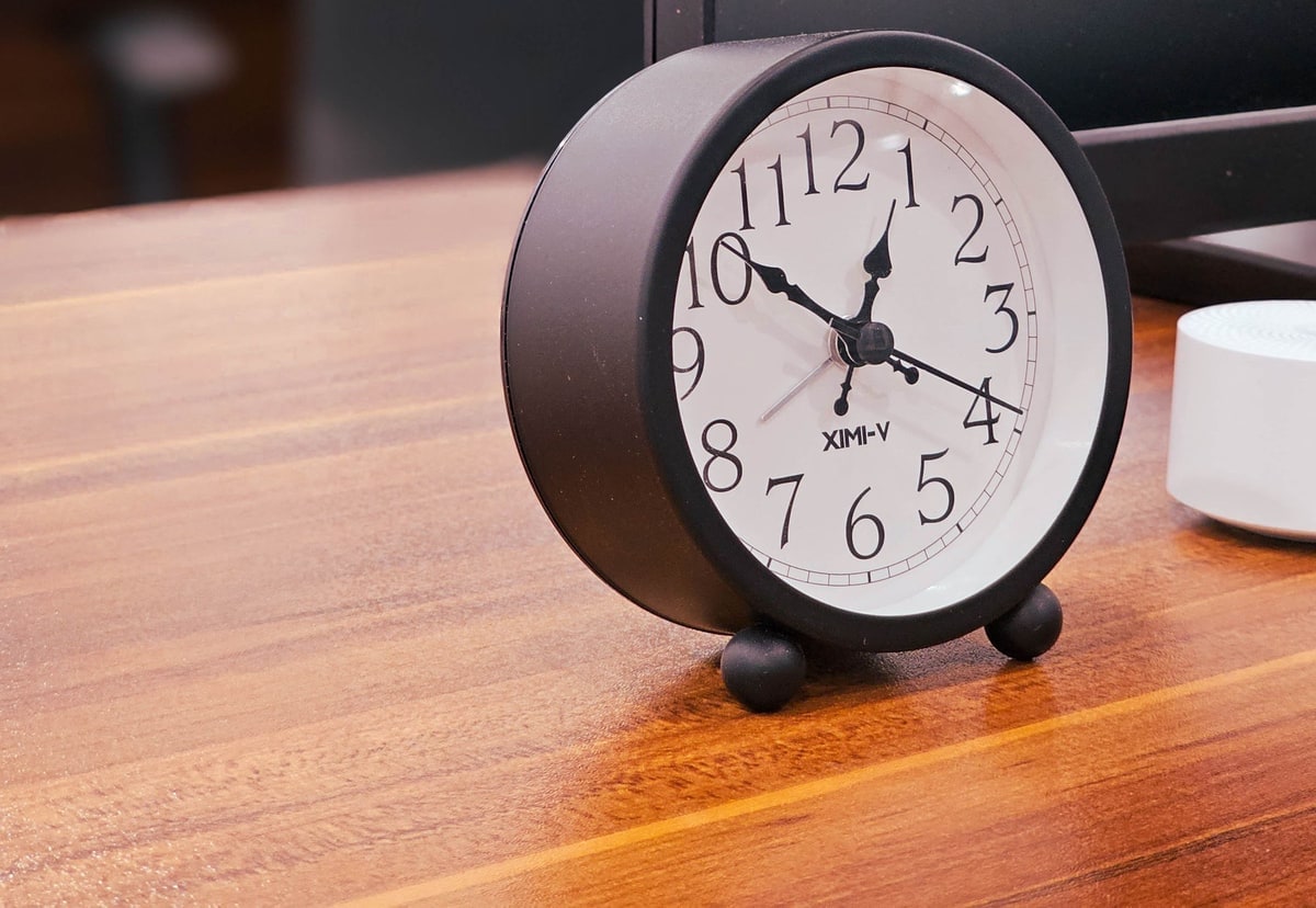 An analogue clock beside a computer monitor on a wooden office desk