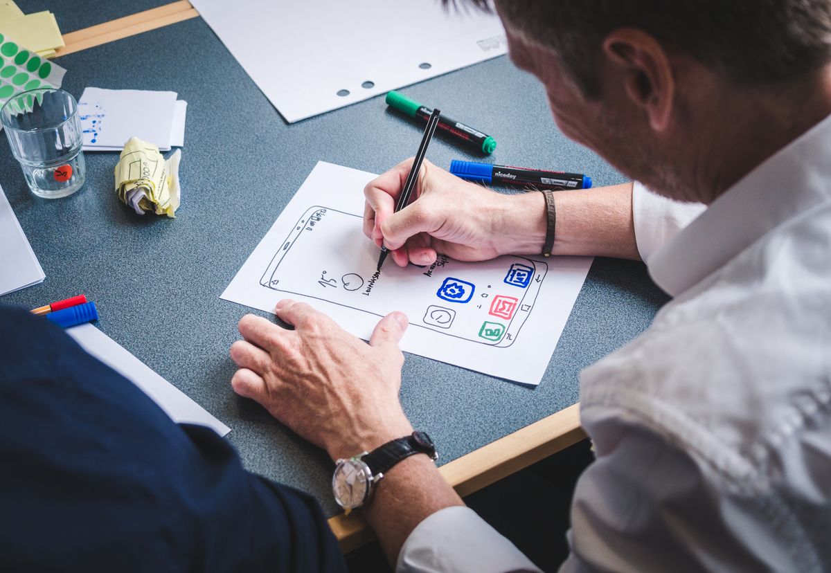 A designer drawing a paper mobile interface prototype at a desk with coloured pens and notes