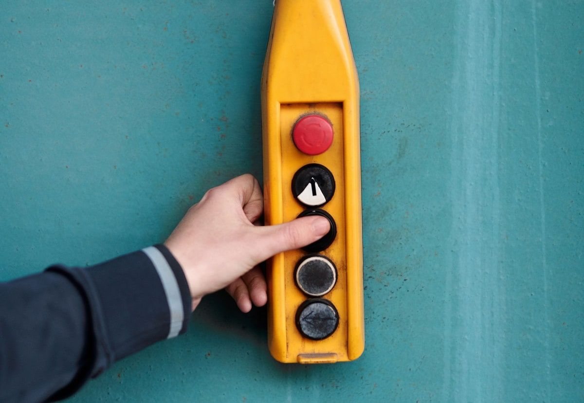 A hand pressing a button on a yellow industrial control pendant against a teal wall