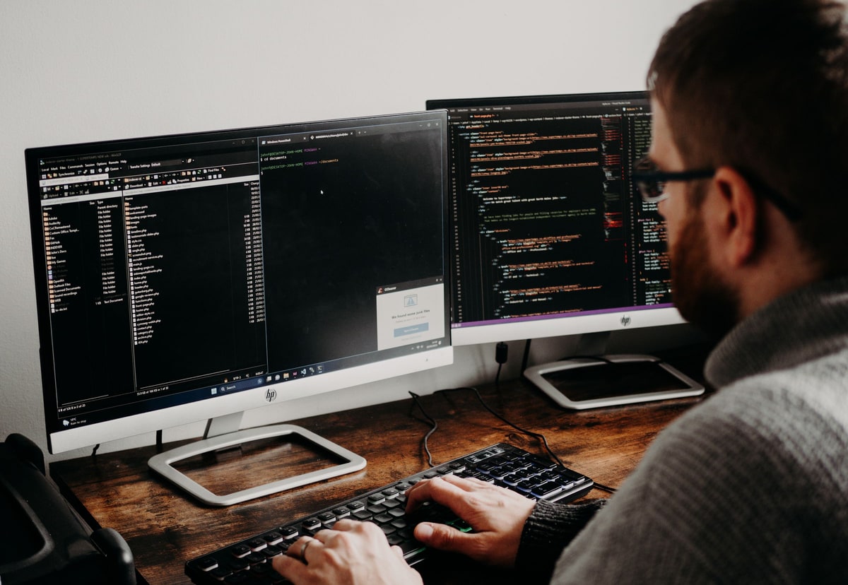 A software developer working across two computer monitors with code and application files visible