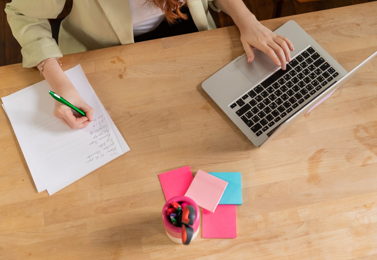 A person writes a process on paper beside a laptop and colourful sticky notes on a wooden desk