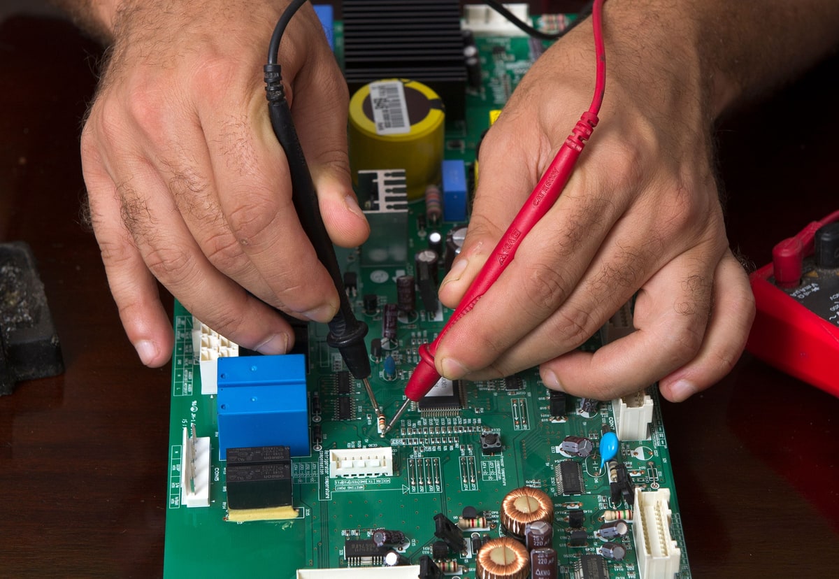 Hands using red and black test probes to check one component on a green circuit board